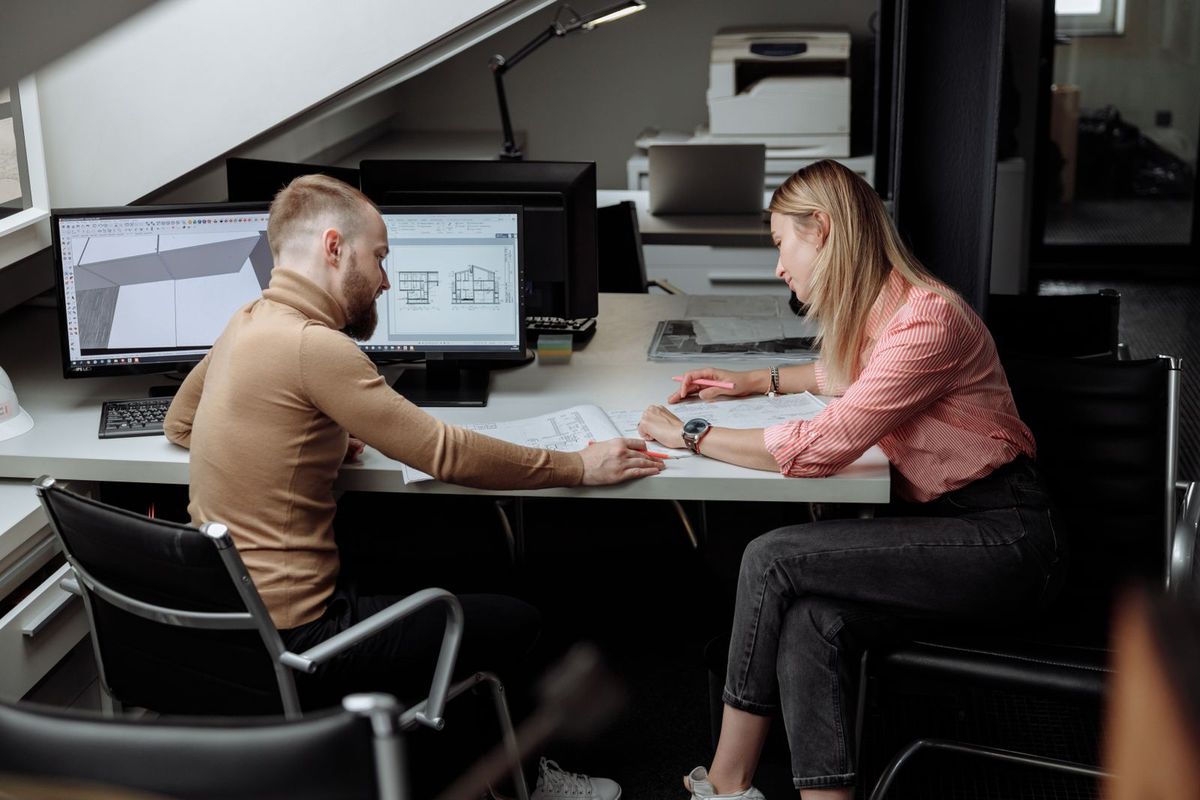 Two colleagues at a shared desk reviewing printed drawings beside monitors showing building plans and sections