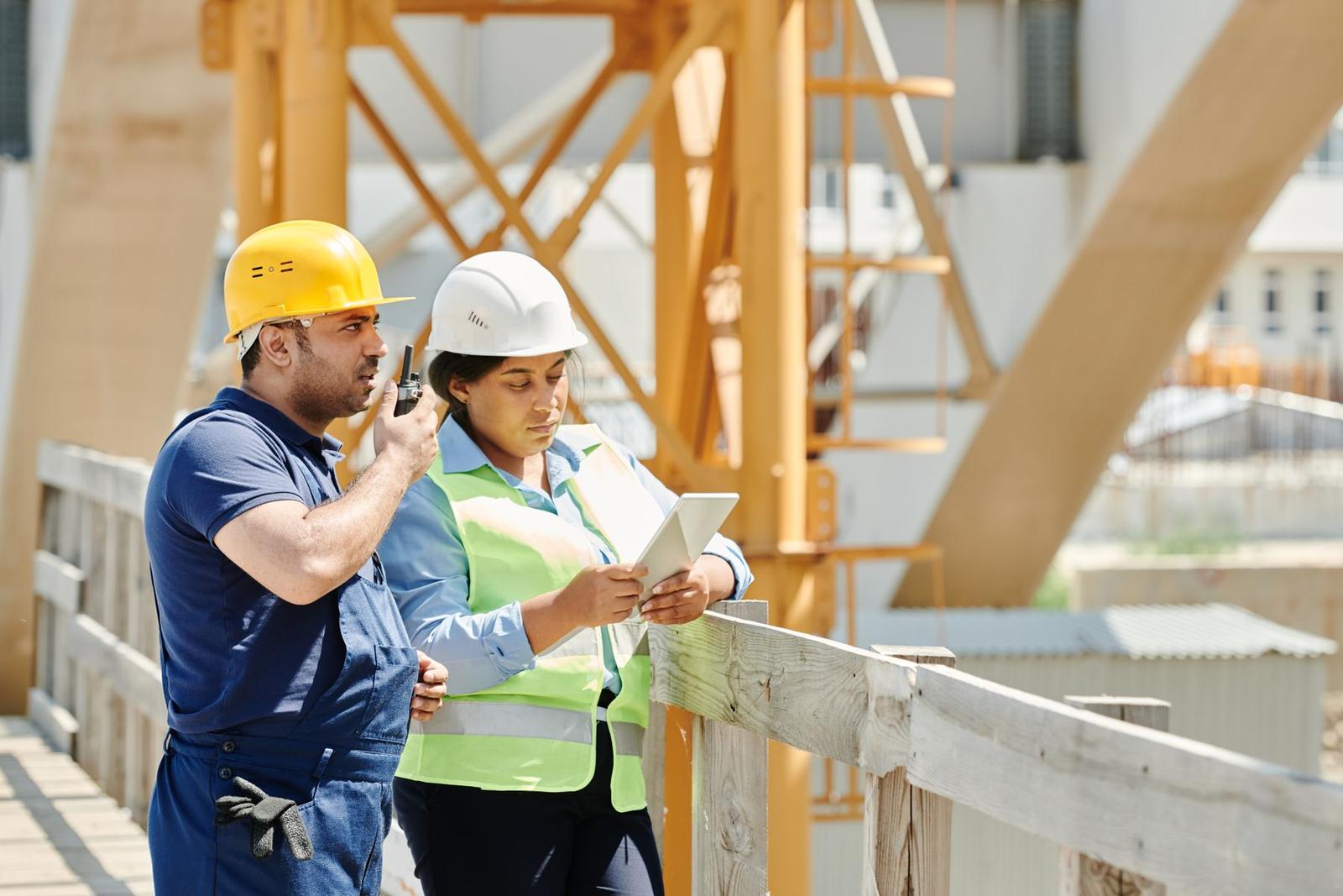 A site manager in a white hard hat and hi-vis checks a tablet while a colleague in a yellow hard hat talks on a radio, on a site walkway beside a tower crane
